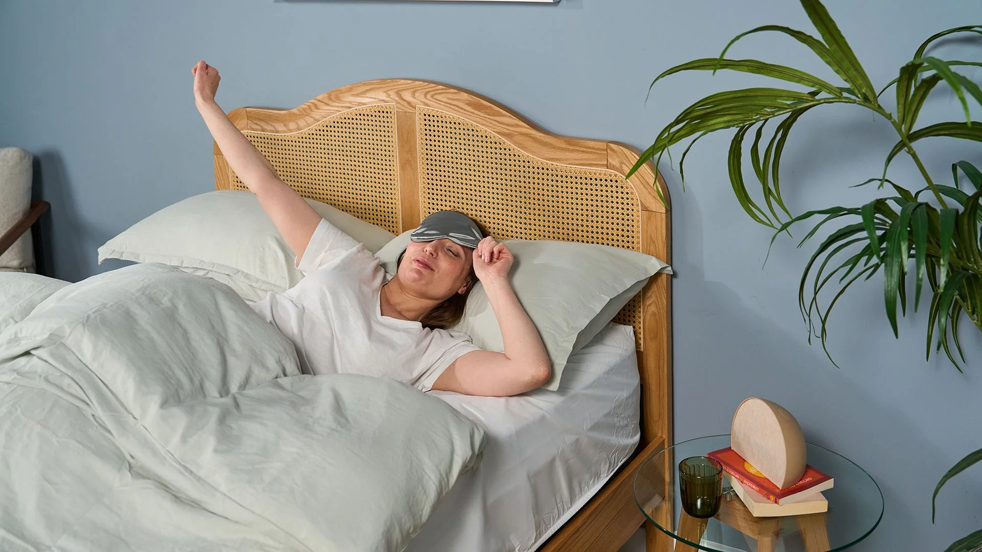 A woman with brown hair and wearing a silver silk eye mask wakes up, stretches and smiles after a great night's sleep