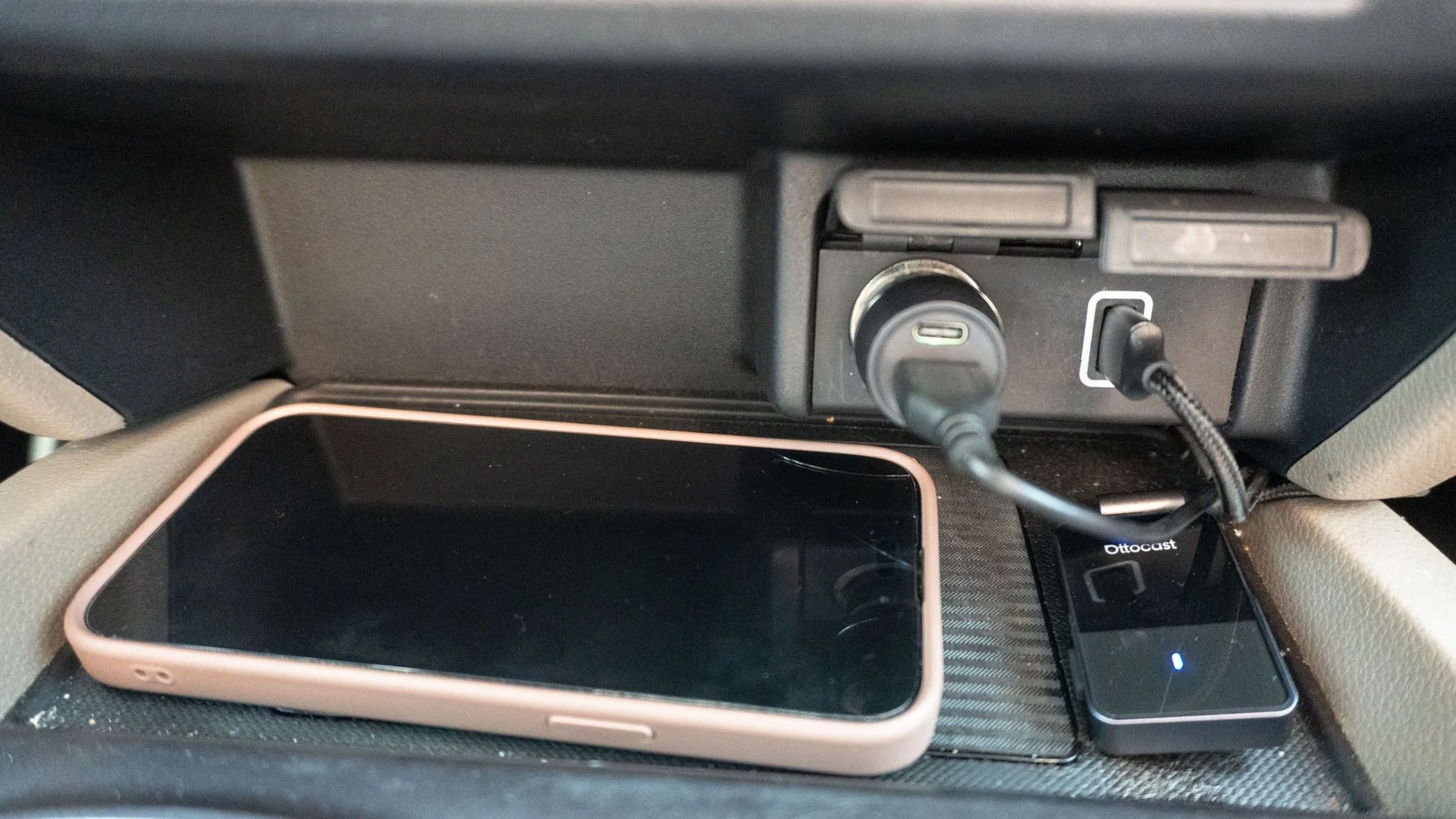 A phone charging on a wireless charging pad next to the Ottocast OttoSafe Cam's wireless adapter plugged into a car's USB port